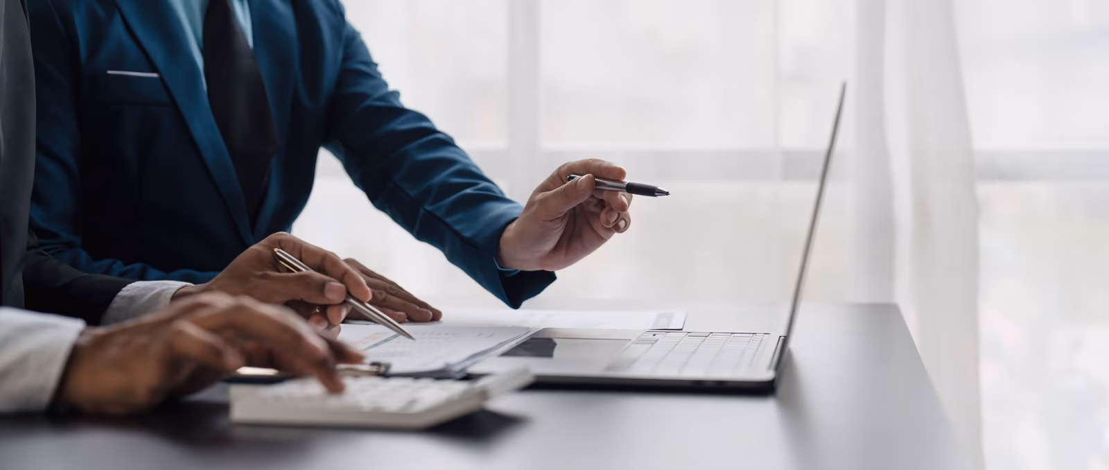 Dos personas en un ambiente corporativo analizan documentos y datos financieros sobre una mesa, utilizando una calculadora y una laptop. Una de ellas señala la pantalla con un bolígrafo, mientras la otra toma notas. La imagen representa un momento de análisis y toma de decisiones, destacando la importancia de la estandarización de datos para garantizar precisión y eficiencia en los procesos empresariales.