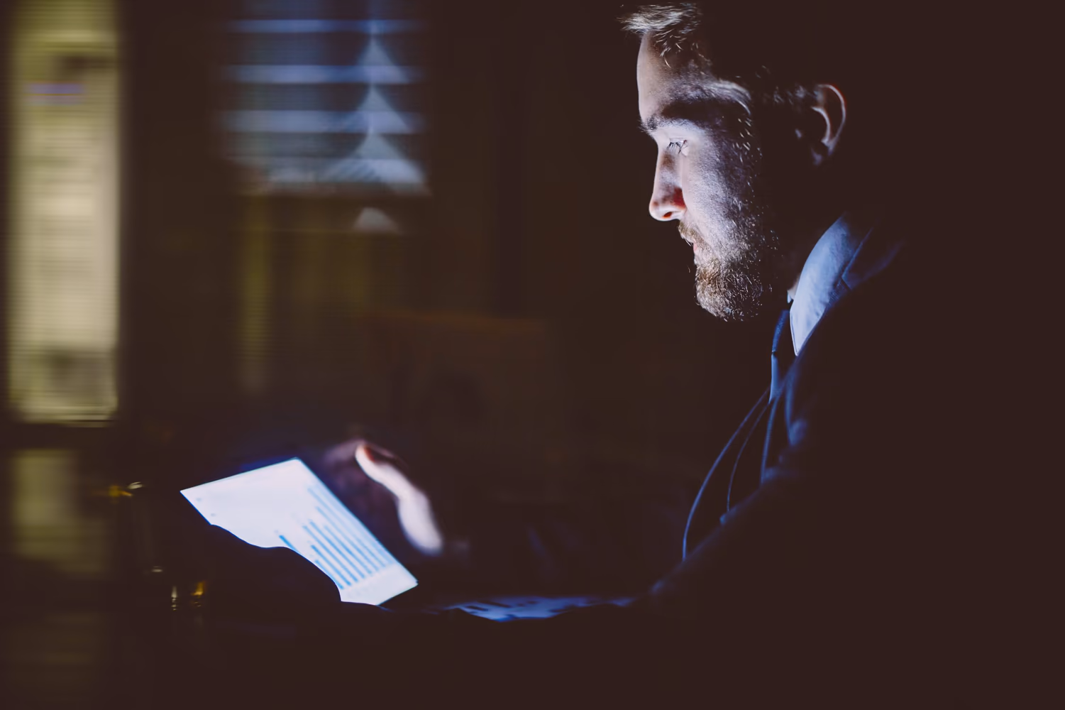 La imagen muestra a un hombre en un ambiente oscuro, vestido con traje, utilizando una tablet que emite una luz intensa. Esta escena puede asociarse al proceso de registro de productos, indicando el uso de tecnología para gestionar información de forma estratégica, automatizada y eficiente, incluso en situaciones que exigen atención detallada y toma de decisiones rápida.