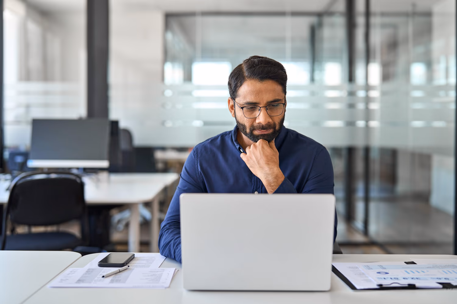 Hombre en un ambiente corporativo se concentra en una laptop mientras analiza información. Sobre la mesa hay documentos, gráficos y un smartphone, indicando una tarea analítica. La imagen transmite el concepto de enfoque y estrategia, representando a un profesional que trabaja con Master Data Management (MDM) para mejorar procesos y tomar decisiones basadas en datos.
