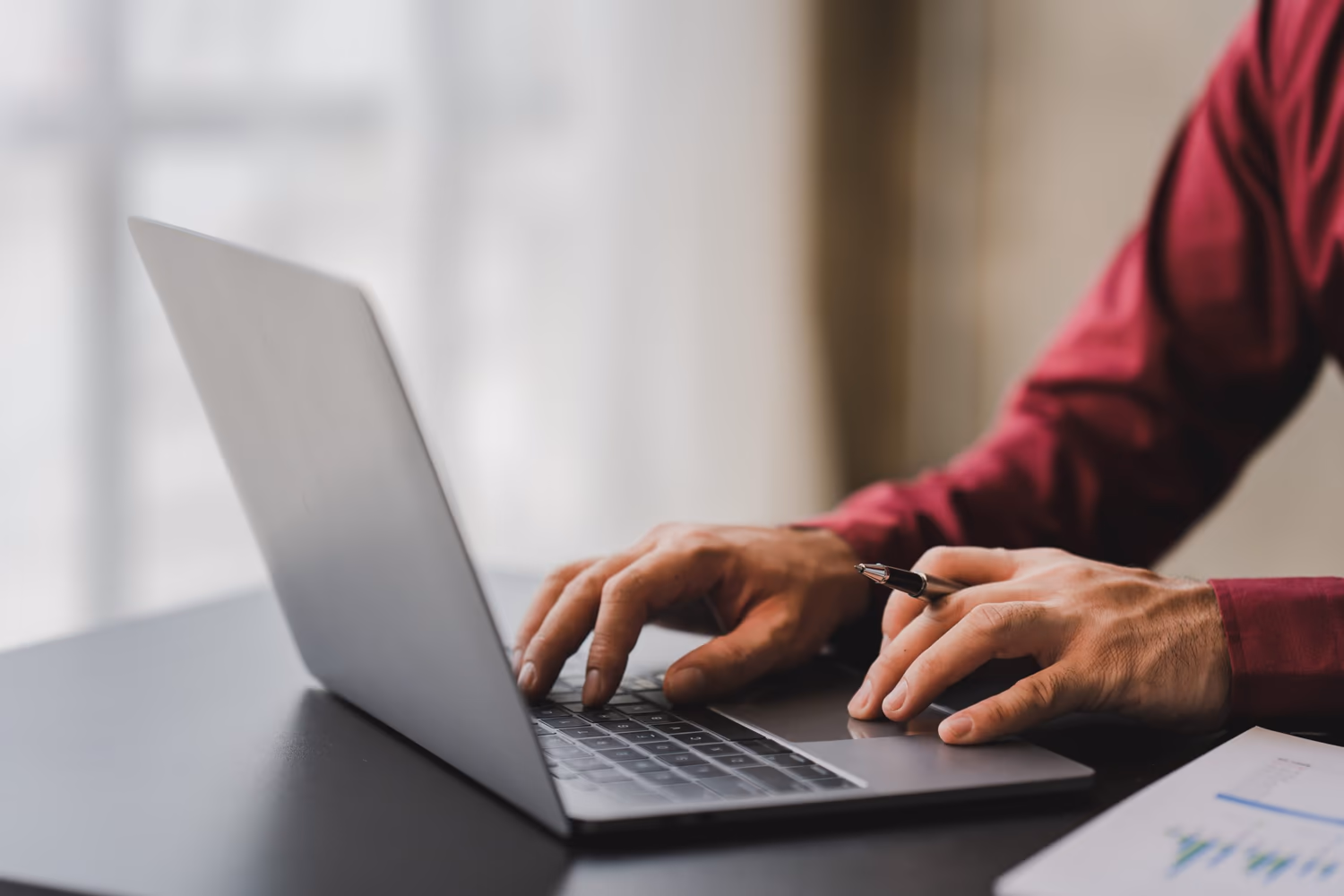 En la imagen, una persona está usando una laptop, con una mano escribiendo y la otra sosteniendo un bolígrafo. Sobre la mesa, al lado de la laptop, hay una hoja con gráficos visibles, lo que sugiere un análisis de datos o una actividad de trabajo relacionada con datos.