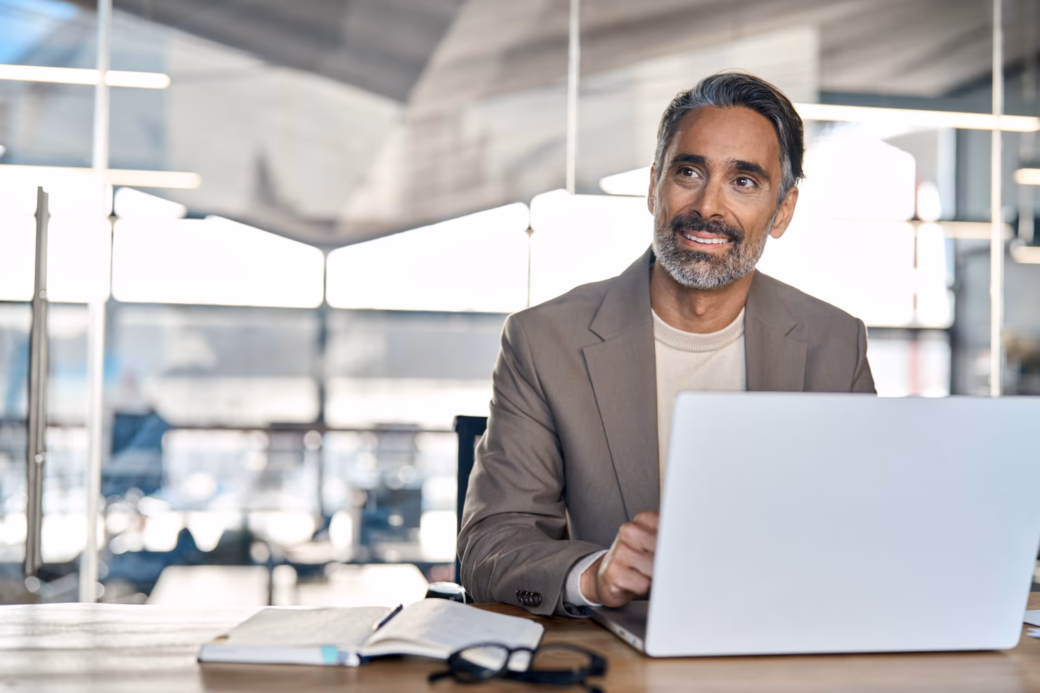 Hombre de mediana edad, con barba canosa y expresión confiada, está sentado en una oficina moderna e iluminada, utilizando una laptop. Viste un blazer beige claro y una camiseta clara. La imagen representa un momento de análisis o toma de decisión, simbolizando el proceso estratégico y riguroso de homologación de proveedores en entornos corporativos.