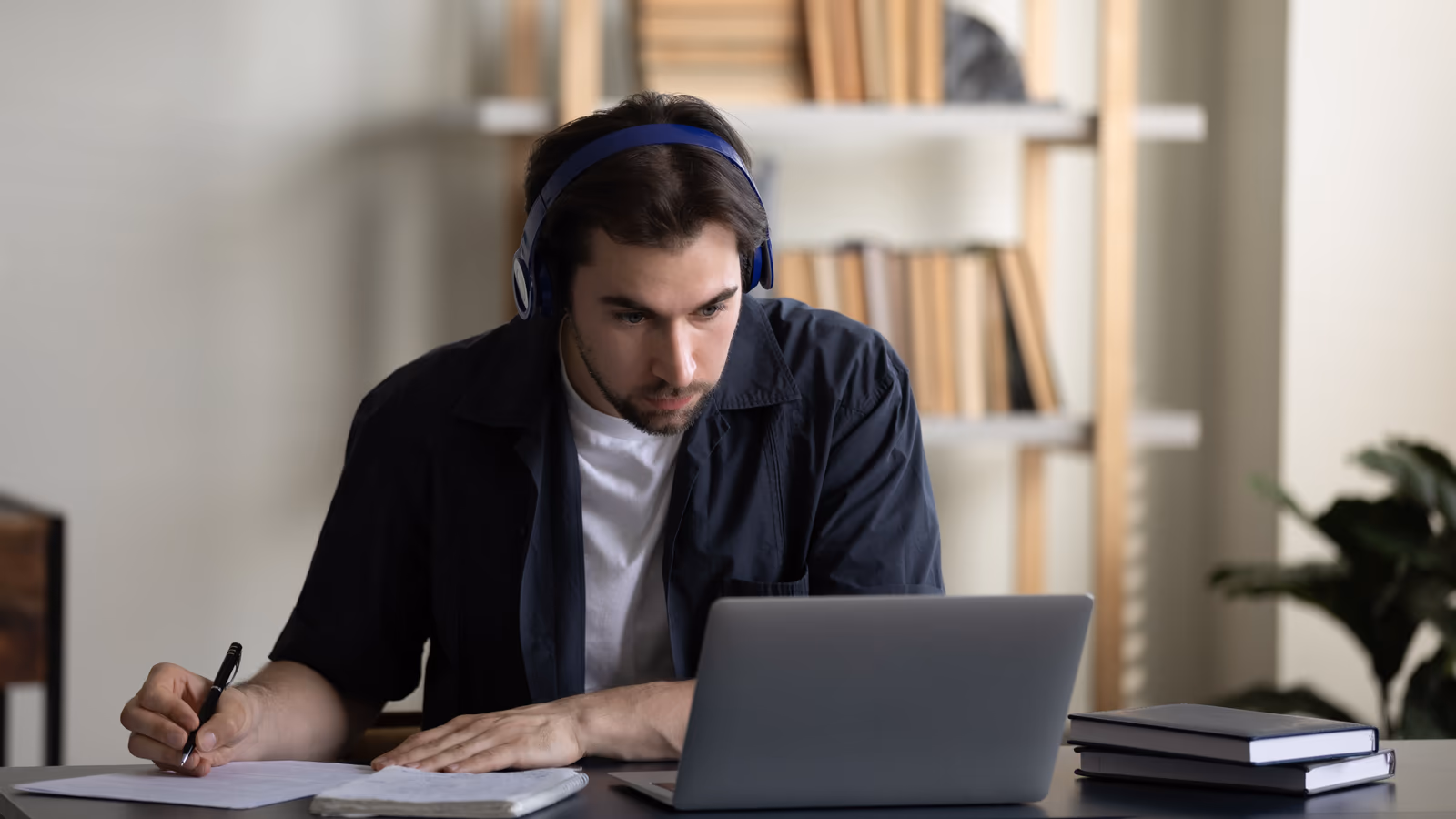 La imagen muestra a un hombre joven concentrado, trabajando en una laptop en un entorno de oficina en casa. Lleva auriculares y mira atentamente la pantalla mientras escribe notas en un papel.