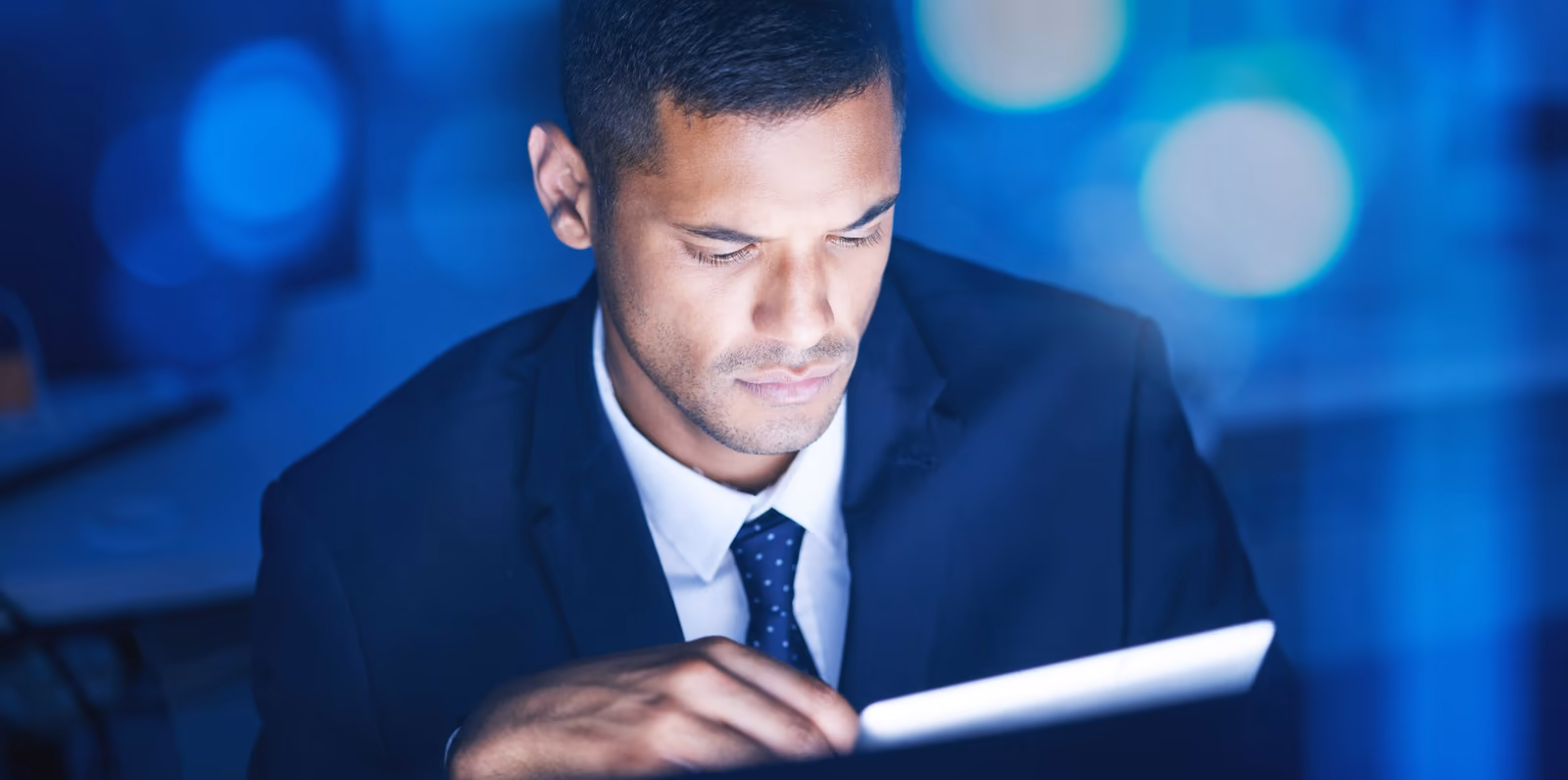 Hombre mirando la pantalla de una tableta, en un ambiente iluminado con tonos azules. La expresión seria transmite concentración, sugiriendo análisis de información o trabajo con base de datos en un contexto profesional y tecnológico.