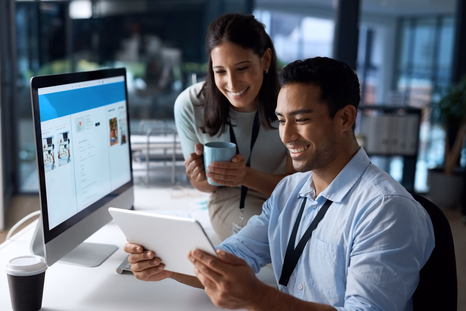 Dos profesionales en una oficina moderna analizan una tableta con sonrisas. El hombre, de camisa azul, está sentado, mientras que la mujer, de pie, sostiene una taza. Sobre la mesa hay una computadora que muestra un sitio web, sugiriendo un trabajo colaborativo, posiblemente vinculado al registro de productos o proveedores.
