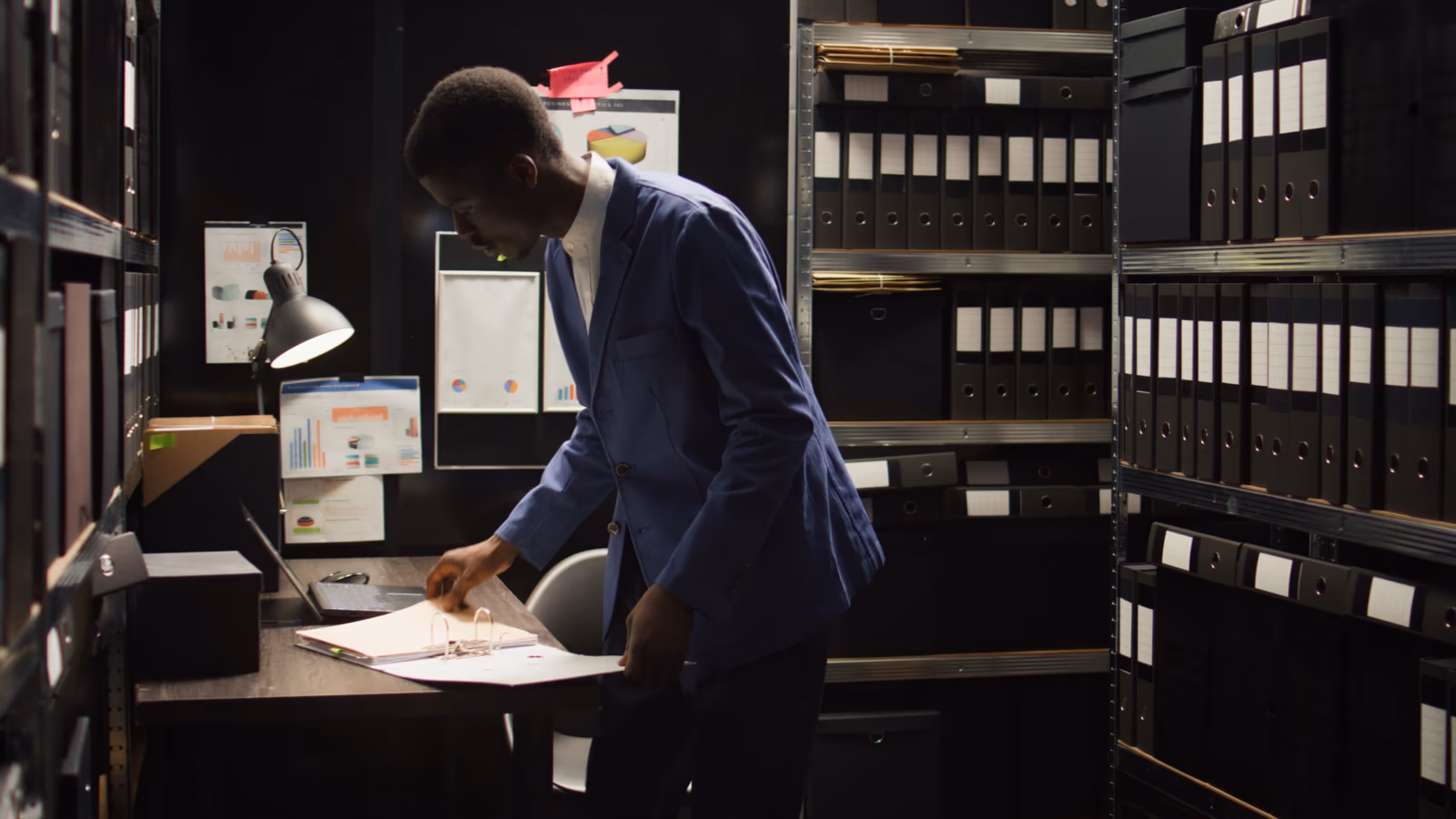 Profesional en un ambiente corporativo, de pie junto a una mesa, analizando documentos en una carpeta. El lugar está rodeado de estanterías con archivos organizados. Gráficos e informes están fijados en la pared del fondo, reforzando el clima de análisis y auditoría. La escena transmite un ambiente de trabajo enfocado en la verificación de información y el control documental, ilustrando la importancia del background check en la toma de decisiones seguras dentro de las empresas.