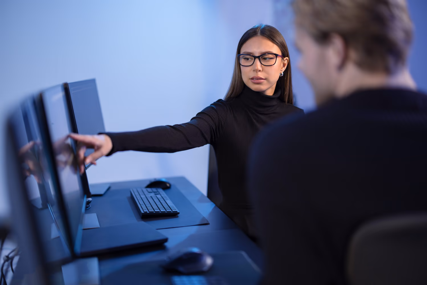 Una mujer con gafas y suéter negro señala la pantalla del computador mientras le explica algo a un colega en un entorno corporativo con múltiples monitores. La imagen representa la gobernanza de datos, evidenciando la colaboración y el análisis estratégico de la información.