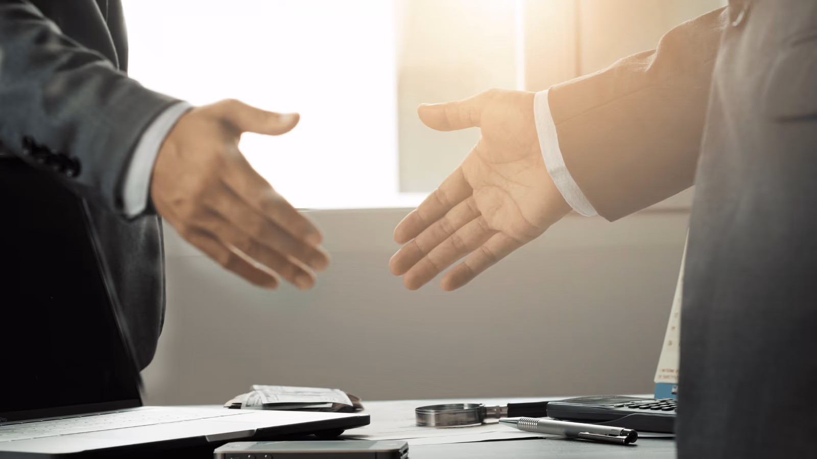 La imagen presenta a dos profesionales que extienden las manos para un apretón de manos sobre una mesa de oficina. Los detalles incluyen la vista parcial de una laptop abierta, algunos papeles y un bolígrafo sobre la mesa, lo que sugiere un ambiente de negocios formal. La iluminación suave contribuye a un clima serio y profesional.