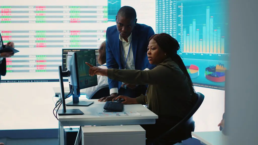 A imagem mostra um ambiente corporativo de análise de dados. Uma mulher sentada aponta para a tela do computador enquanto um homem ao lado observa atentamente. Ao fundo, painéis digitais exibem gráficos e tabelas, sugerindo um cenário de governança de dados e MDM (Master Data Management).