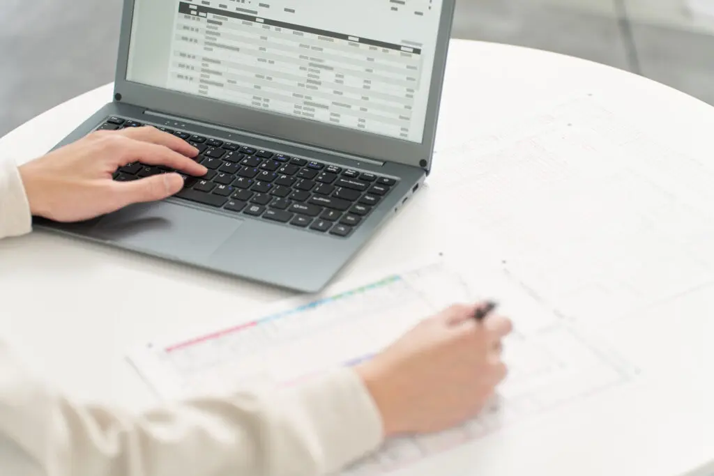 Pessoa trabalhando em uma planilha no computador, com documentos físicos à frente, representando o processo de gestão de dados de forma manual.